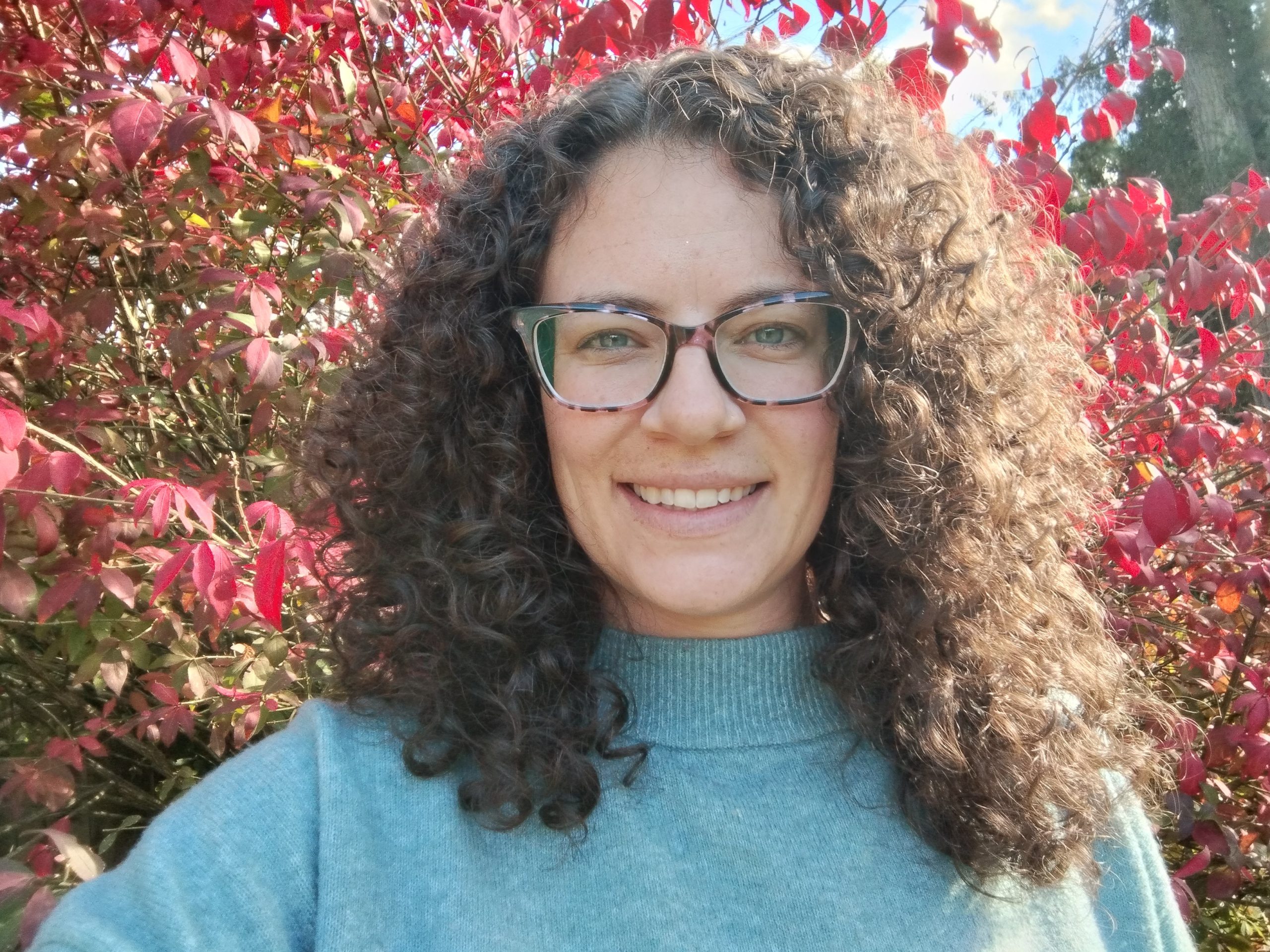 A white woman stands in front of a tree full of red leaves. She has long curly hair and glasses and is smiling at the camera. She is wearing a blue sweater. 